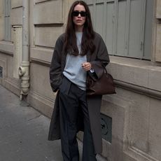 Annabel wears a long brown coat, gray t-shirt, gray trousers, and a brown leather bag.